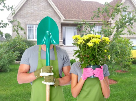 A person and a child wearing green gardening aprons and gloves are standing on a well-maintained front lawn of a suburban house with beige brick walls, white window frames, and a pitched roof. The adult is holding a green shovel with a shiny metal blade, while the child is holding a pink container filled with bright yellow flowers. The garden features a lush, green grass lawn, a mulched flower bed with small shrubs, and a young tree with fresh green leaves in the background. The scene is set during daytime in clear weather, with soft natural lighting emphasizing the vibrant colours of the flowers and greenery. This outdoor space showcases typical residential garden elements, suitable for landscaping and gardening maintenance services provided by Gardening Services Shepherds Bush, designed to enhance curb appeal and outdoor enjoyment in the Shepherds Bush area, W12 postcode.