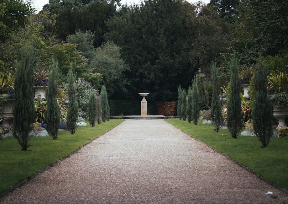 A person wearing a blue raincoat with the hood up, standing on a neatly maintained grassy lawn in a garden, engaged in garden maintenance by using a hand rake to gather fallen leaves. The garden featu